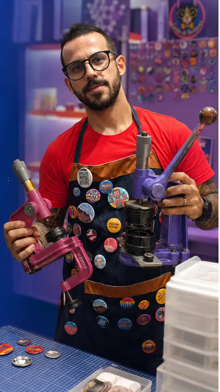 Henrique Maiani na produção da Escola de Bottons, segurando duas prensas de bottons