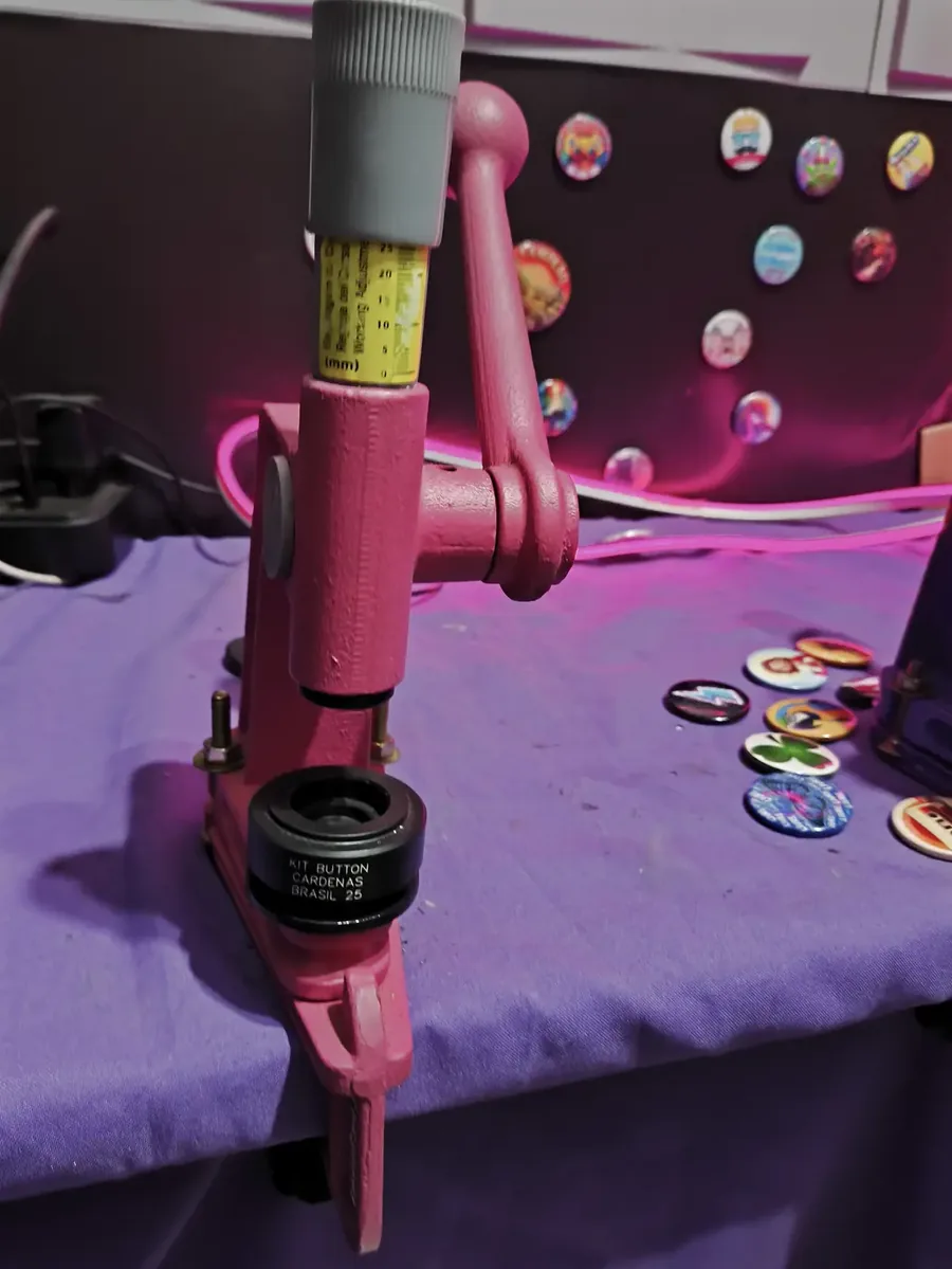 Prensa manual de bottons rosa sobre a bancada, com bottons prontos ao lado